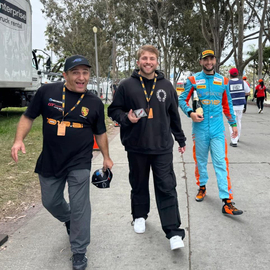 Zia Shlaimoun walks with his sons Nico and Ricco at a motorsport event; Nico wears a black hoodie while Ricco, dressed in a bright racing suit and holding a bottle, celebrates alongside his father