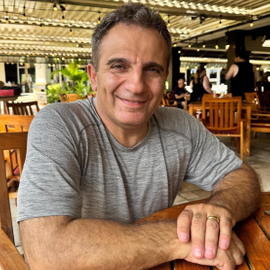Zia Shlaimoun smiles in an open‑air restaurant, dressed in a gray t‑shirt with string lights and patio seating in the background