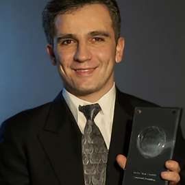 Zia Shlaimoun smiles in a formal suit and patterned tie while proudly holding a Barclays Bank trophy against a dark backdrop