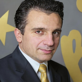 Professional head‑and‑shoulders portrait of Zia Shlaimoun in a dark suit with a golden tie, posed against an abstract gold graphic background at a BIC Enfield event.